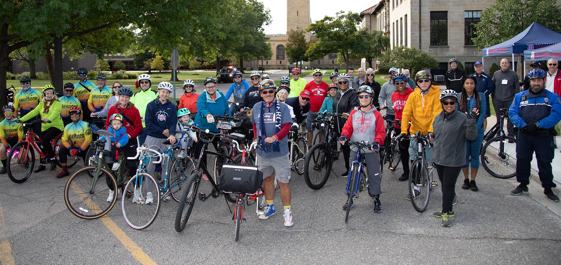More than three dozen people, many with bikes stand outside in front of buildings, trees and a clock tower.