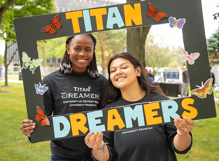 two students posing with titan dreams prop sign by fountain