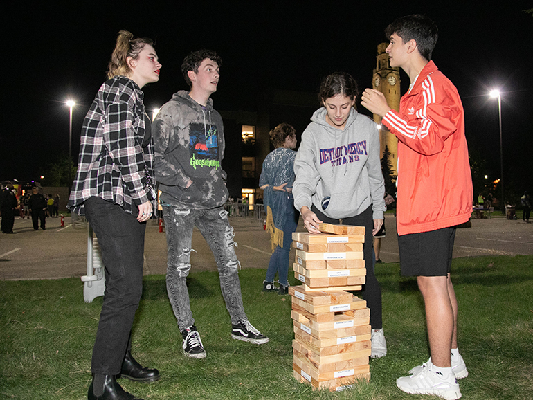 Students socialize and play a giant Jenga game during Homecoming. The clocktower is visible and lit up in the background.