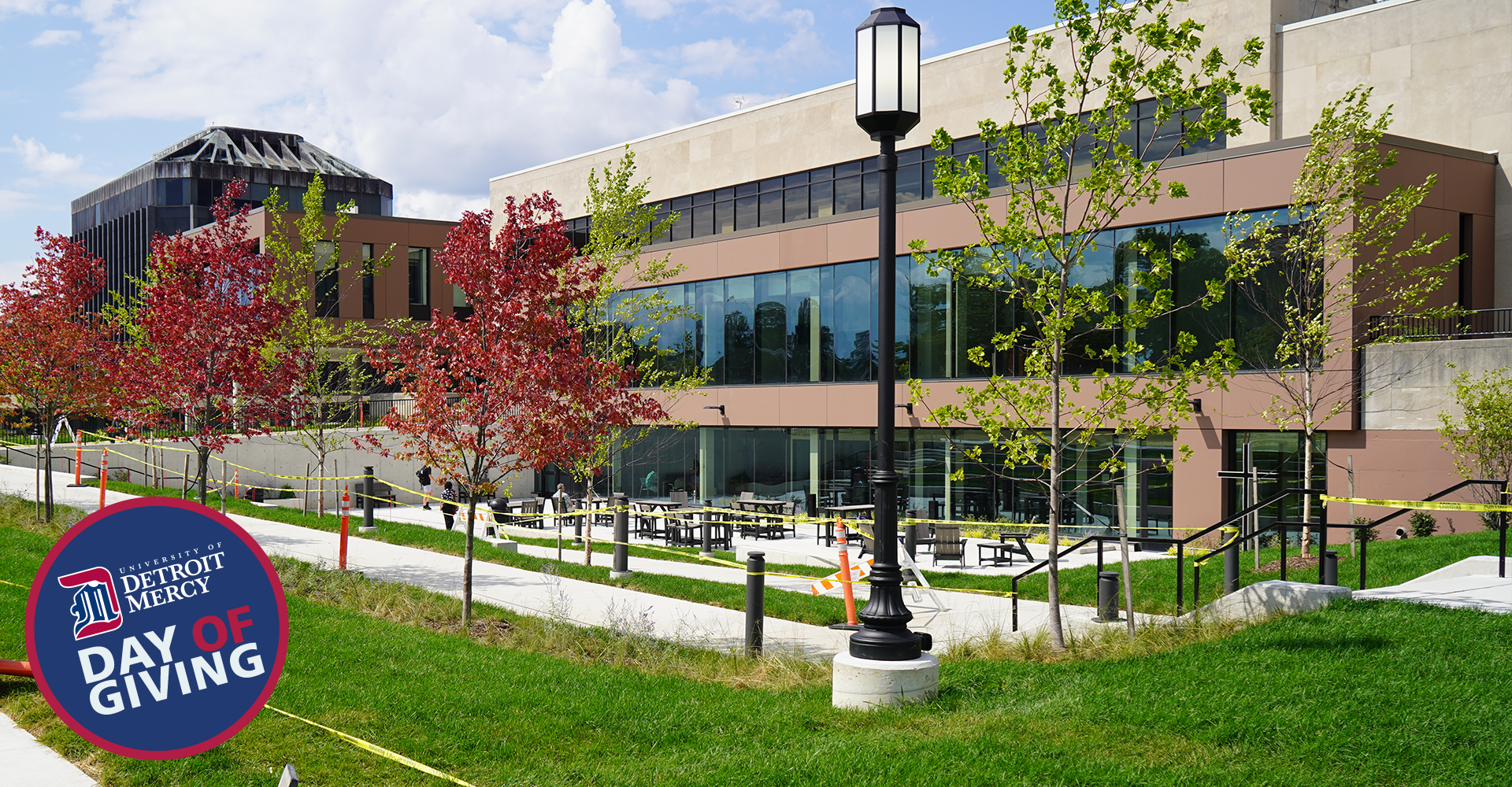 An outdoor photo of the Student Union plaza with trees and lamp posts in front. A University of Detroit Mercy Day of Giving sticker is in the lower left hand corner.
