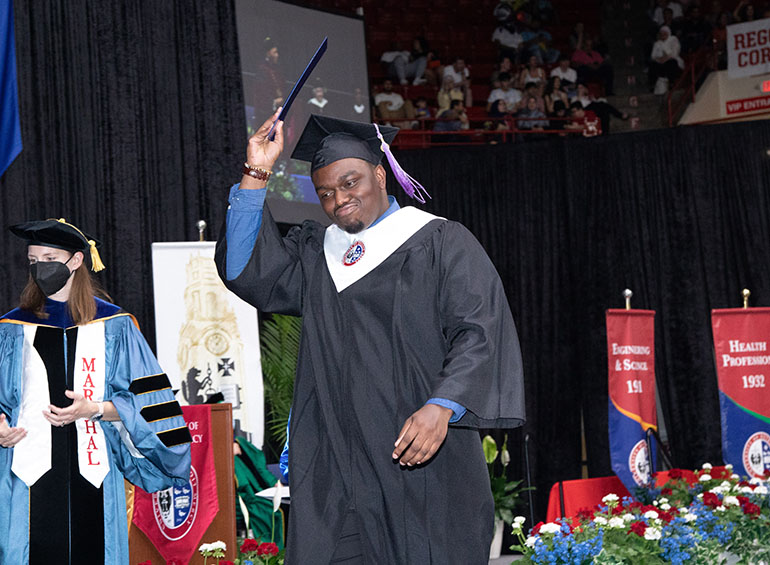 A graduate smirks and lifts his diploma folder up in celebration while walking on stage during commencement.