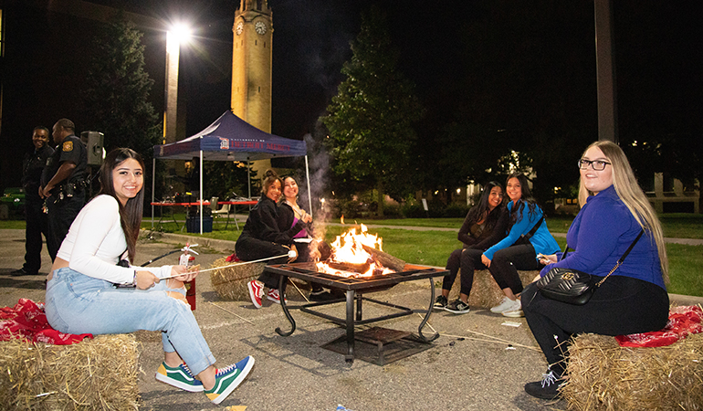 Six people sit on bales of hay outdoors making s'mores over a fire. A clock tower, trees and buildings are pictured in the background.
