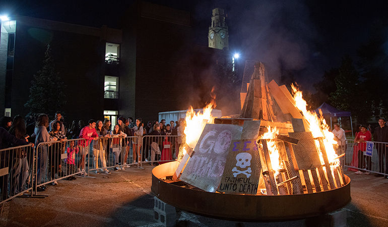 A large bonfire with signs in it sits in a parking lot outdoors with students surrounding it separated by a fence.