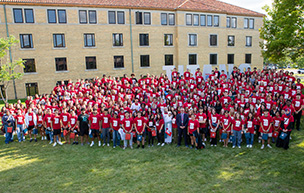 Dozens of people wearing red UDM t-shirts and others stand and pose for a Class of 2028 photo outdoors in front of Holden Hall and the TITANS signage.