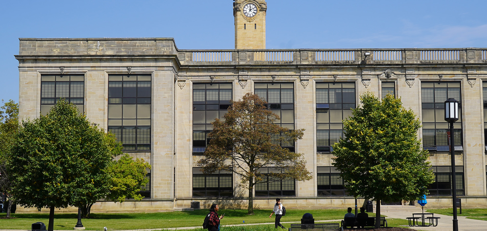 Students walk on the sidewalk in front of the Engineering Building during a summer day, with trees in the foreground and a clock tower in the background.