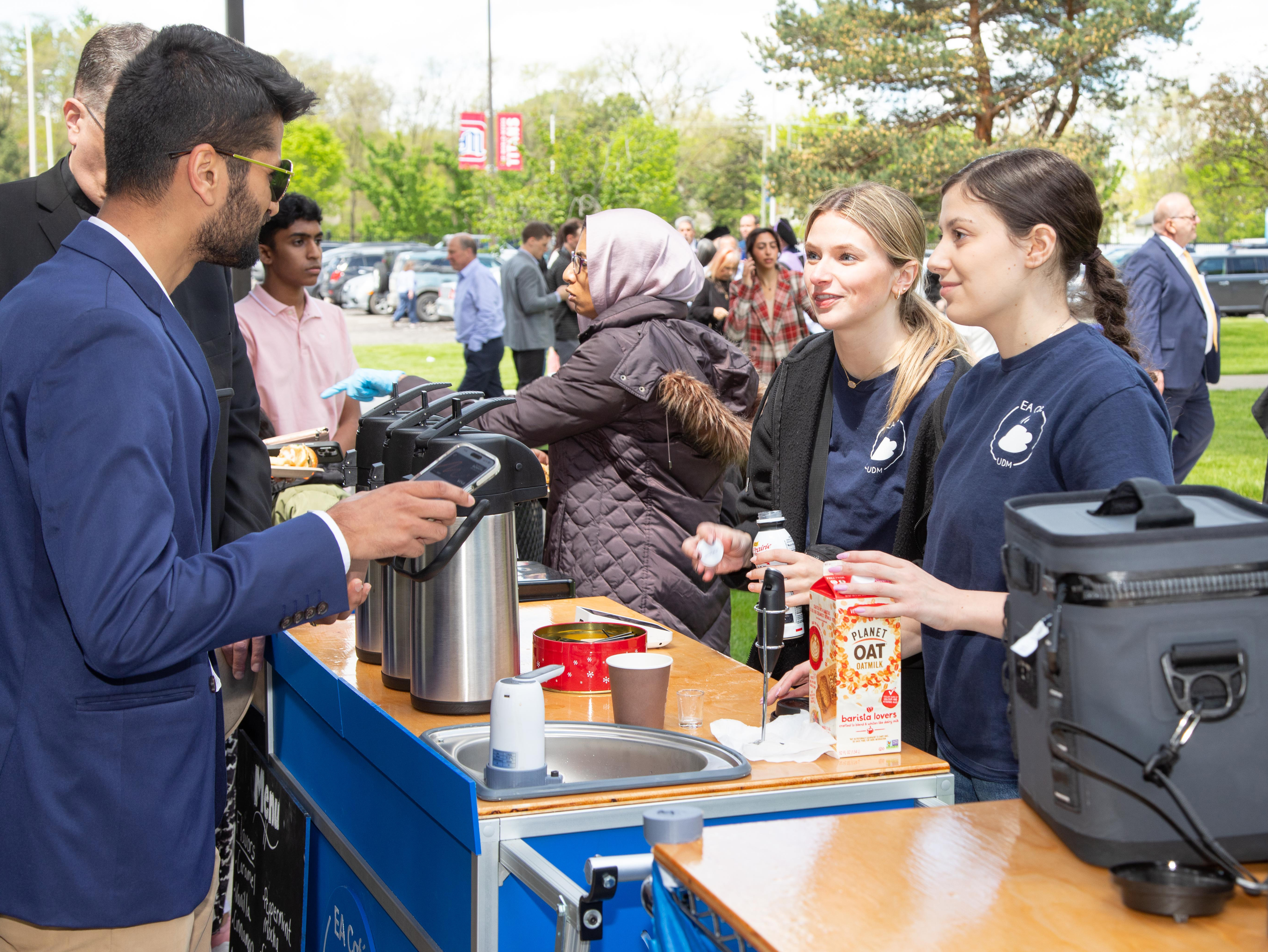 Two students stand near a coffee cart and serve to customers outdoors during commencement.