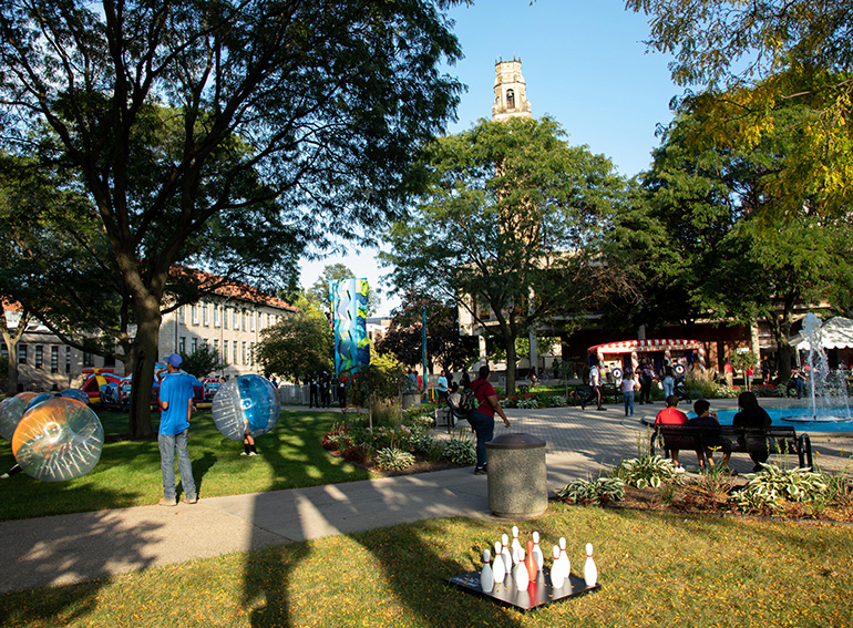 Activities and games are pictured with people outdoors on the McNichols Campus with trees, a fountain, buildings and a clock tower also pictured on a sunny day.