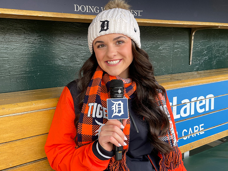 Kara Wolfbauer wearing Detroit Tigers gear sits inside of a dugout at Comerica Park, holding a microphone.