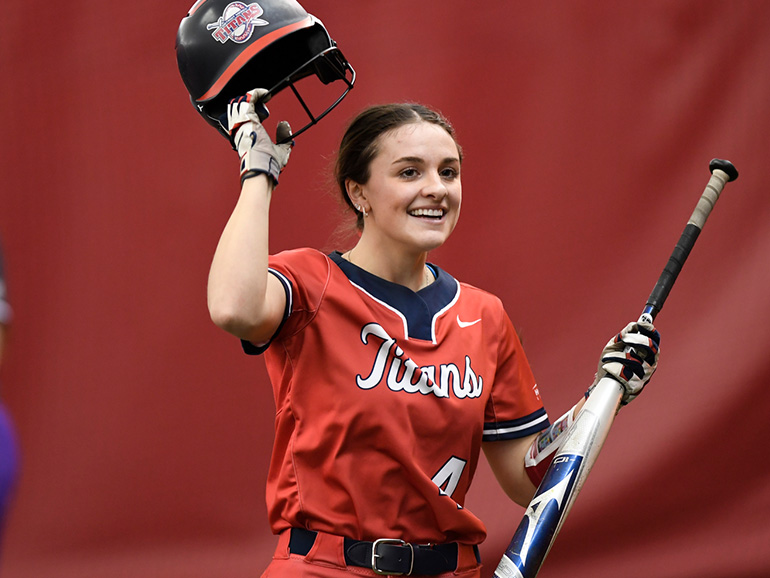 Kara Wolfbauer holds a bat and helmet, while wearing a softball uniform inside of a dome.