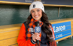 Kara Wolfbauer, wearing Tigers gear, holds a microphone inside of the dugout at Comerica Park.