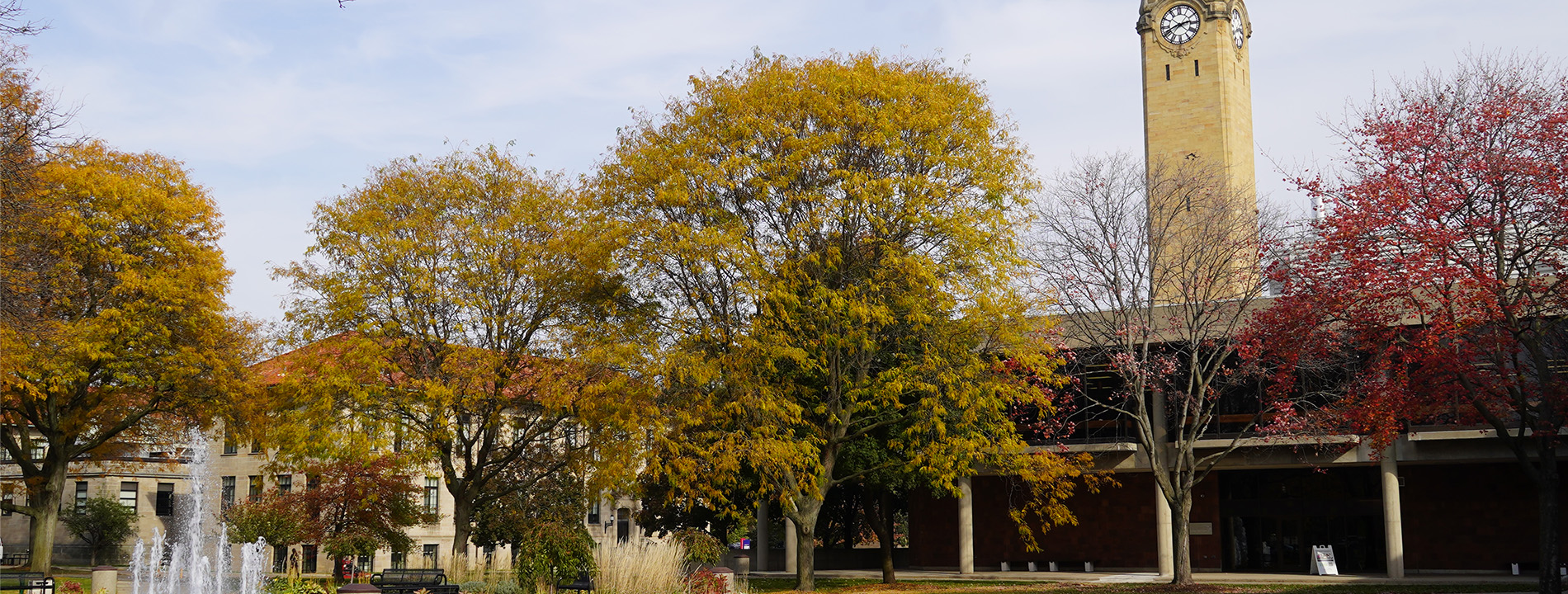 Colorful leaves fill the trees near the Fisher Fountain and Student Union, with a clock tower in the background outdoors.