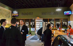 Eight people stand and mingle inside of Boost Detroit with photos, television screens and a countertop pictured in the photo. The words DETROIT BOOST are displayed on the wall.