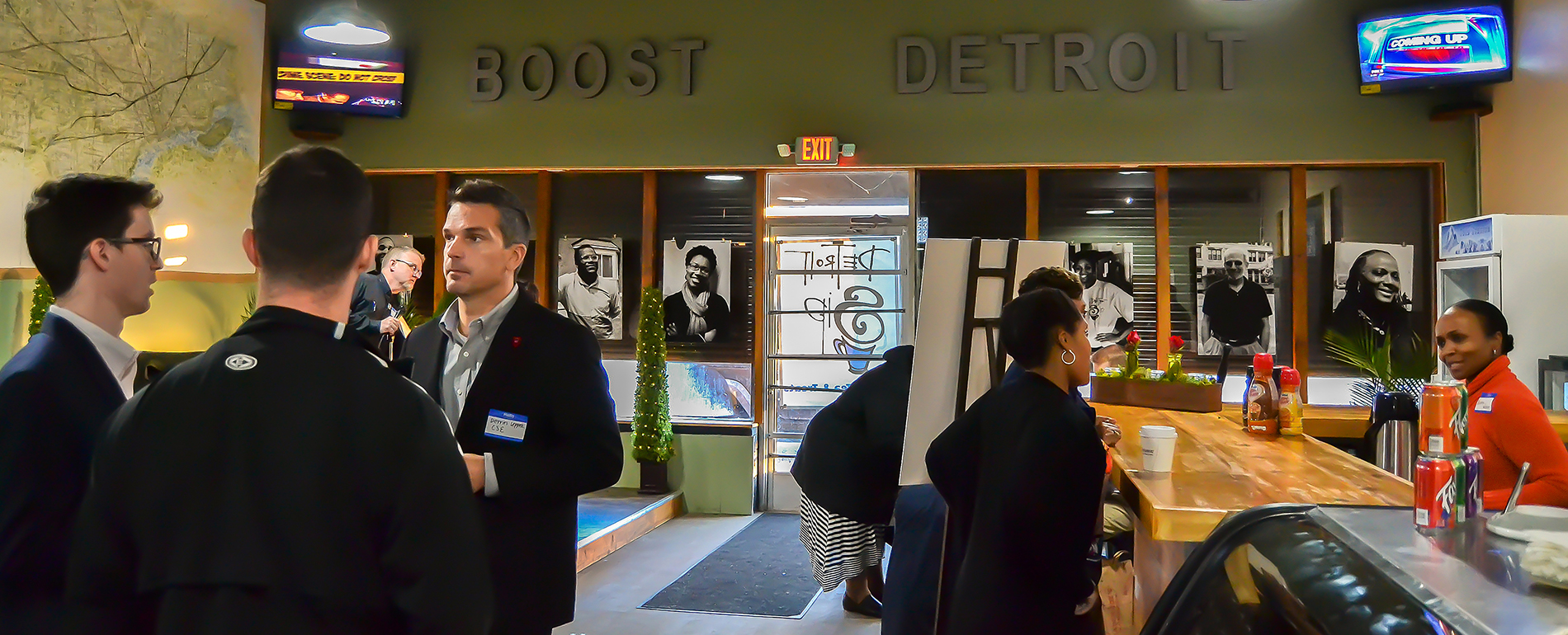 Eight people stand and mingle inside of Boost Detroit with photos, television screens and a countertop pictured in the photo. The words DETROIT BOOST are displayed on the wall.