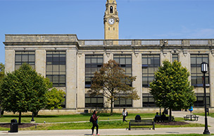 Students walk on the sidewalk in front of the Engineering Building during a summer day, with trees in the foreground and a clock tower in the background.