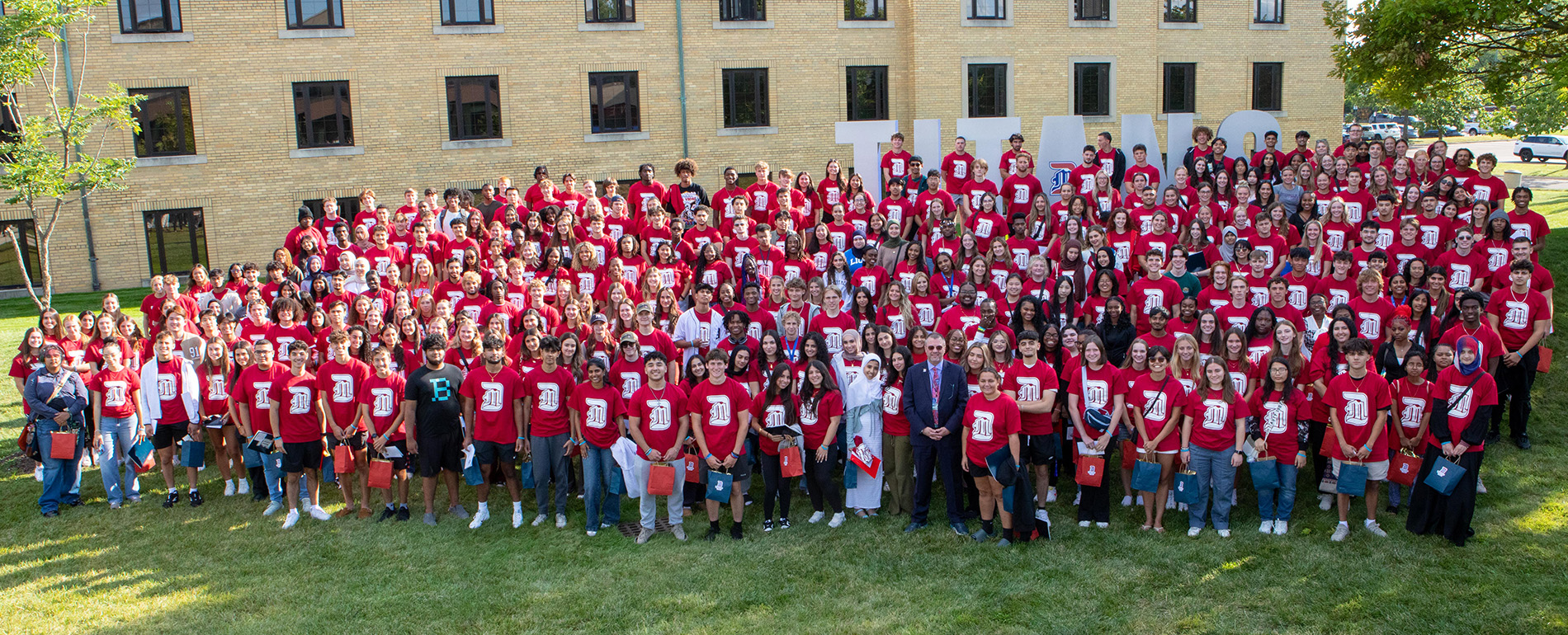 Dozens of people wearing red UDM t-shirts and others stand and pose for a Class of 2028 photo outdoors in front of Holden Hall and the TITANS signage.