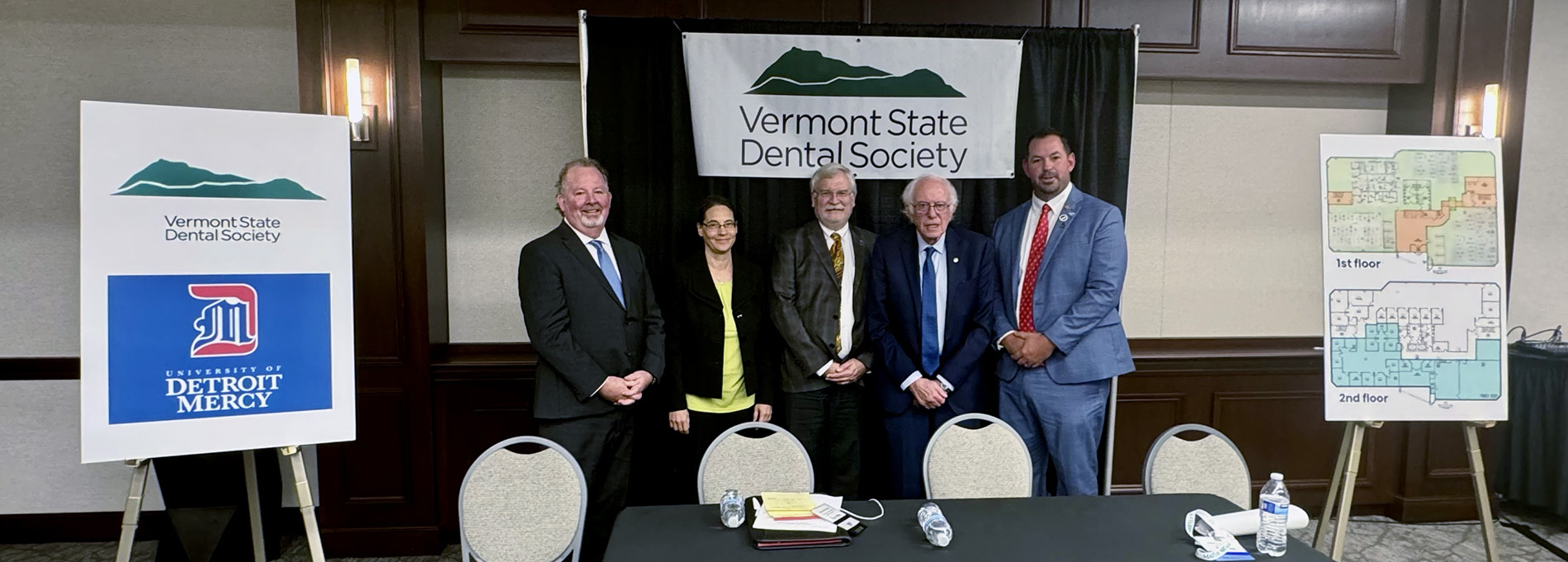 A group of people, including Detroit Mercy School of Dentistry Dean Mert N. Aksu and Sen. Bernie Sanders, pose for a photograph at a press conference announcing a partnership between Vermont State Dental Society and University of Detroit Mercy. Logos and renderings are shown at each end of the table.