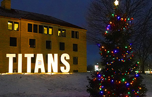 A Christmas tree with colorful lights shines on the McNichols Campus, with TITANS lettering aglow and Holden Hall in the background.