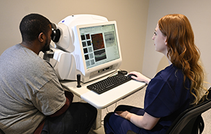 A man sits at a machine during an eye exam while a woman looks at images of the eye.