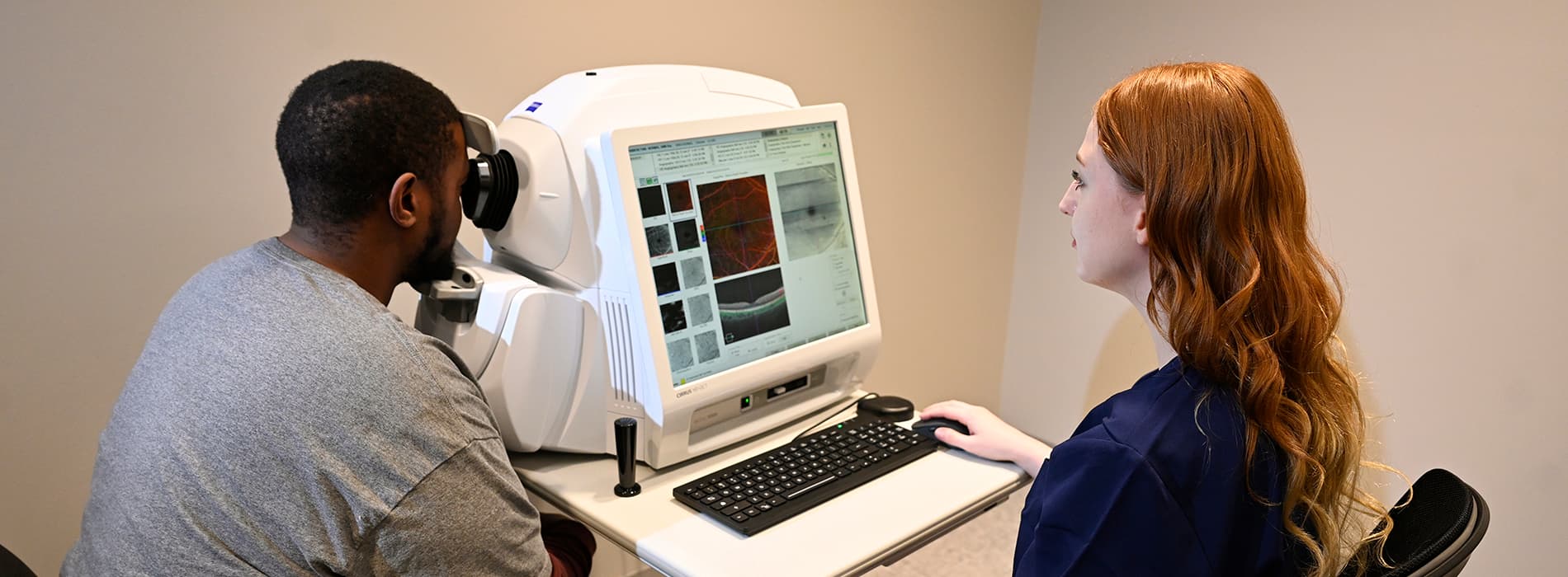 A man sits at a machine during an eye exam while a woman looks at images of the eye.