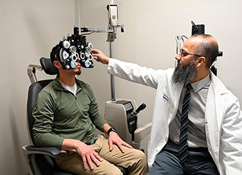 A man sits in a chair while a man uses a phoropter during an eye examination.