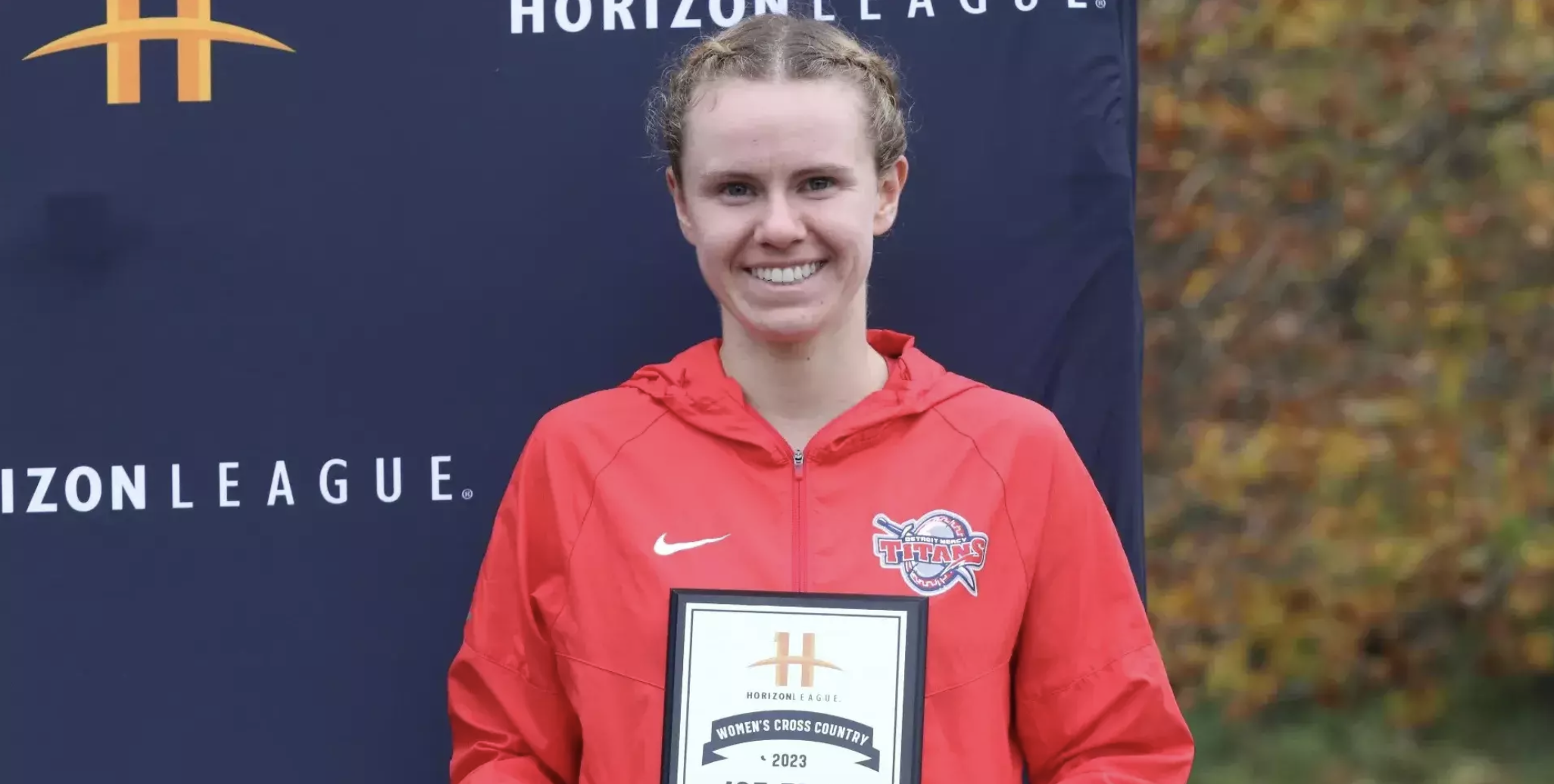 Allison Sherman holds a 2023 cross country plaque outdoors at a championship meet, standing in front of a Horizon League banner.