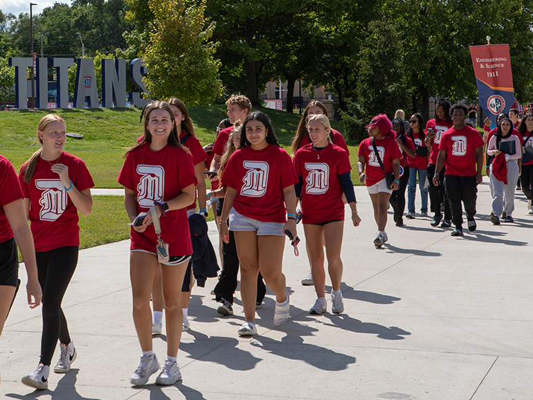 Students wearing red UDM t-shirts walk down the sidewalk outdoors during first-year convocation, with Holden Hall, trees and the large TITANS signage in the background.