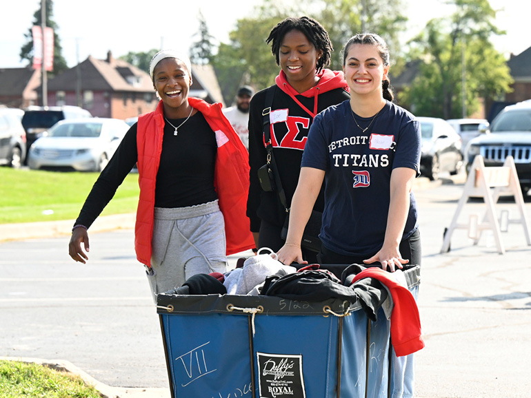 Three students smile and push a trolley outdoors on the McNichols Campus with other people and cars in the background. One student wears a Detroit Mercy Titans t-shirt.