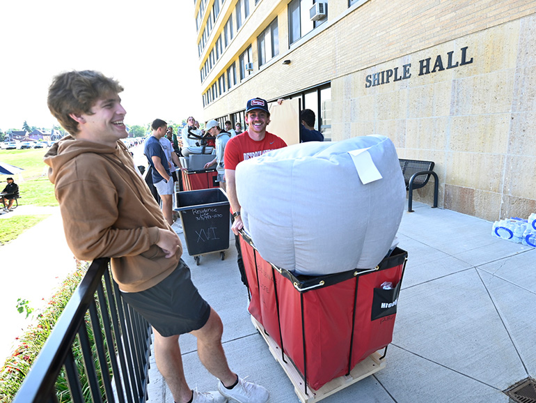 More than a half dozen people stand and wait with belongings outdoors near the Shiple Hall entrance.