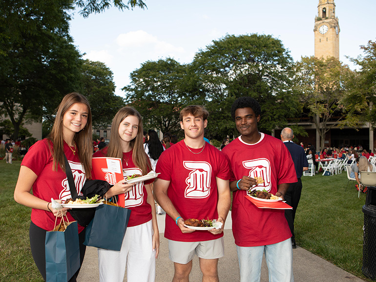 Four students wearing red UDM t-shirts smile and pose for a photo outdoors in front of the Student Union and clock tower.