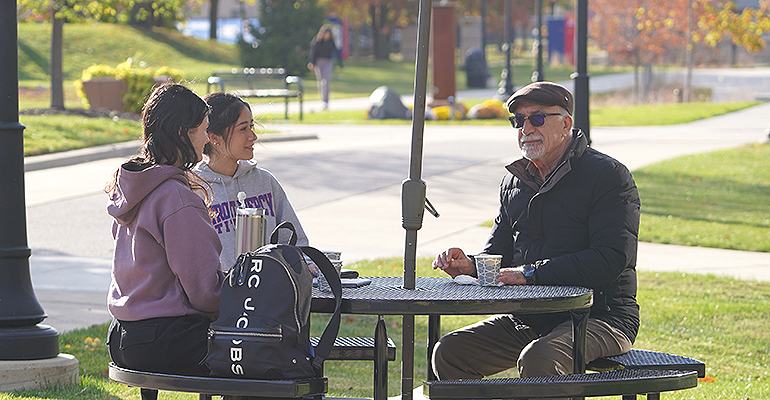 two students sitting at an outdoor table with a community member