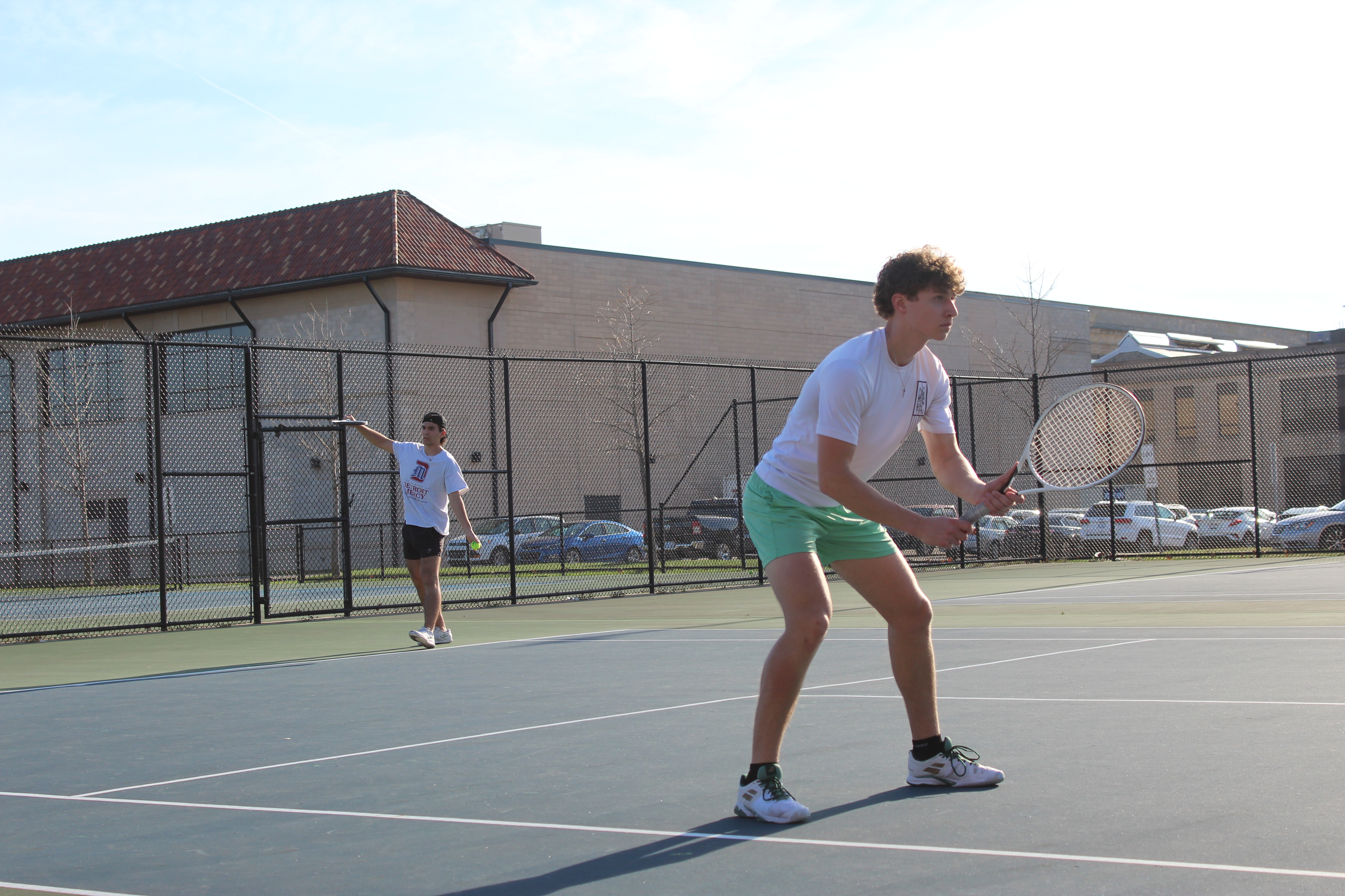 Two students play tennis outdoors on courts with buildings and cars behind them.