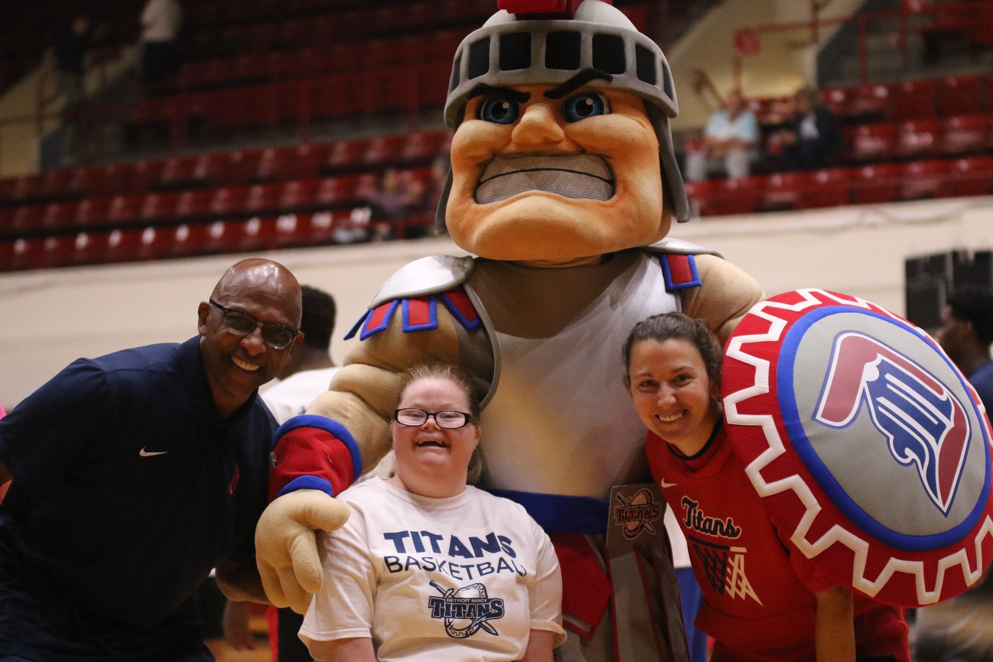 Three people stand inside of Calihan Hall with the UDM mascot, Tommy Titan.